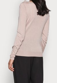 Pull long rose clair à manches longues avec col et poignets côtelés, design ajusté et texture lisse, associé à un pantalon large noir.
