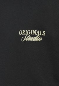 Μαύρο ύφασμα με λευκή κεντημένη επιγραφή "ORIGINALS Studio" στο κέντρο της εικόνας.