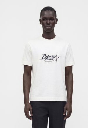 Homme debout face à l'avant portant une chemise blanche à manches courtes avec le texte "Emporio Armani Music District" et un graphisme d'étoile sur le devant.