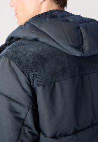 Σκούρο μπλε ζακέτα πλεξίματος με πάνελ ώμου από ταφτά. Διαθέτει μαξιλαρωτή υφή, κουκούλα και λεία ύφανση για ζεστασιά και άνεση.