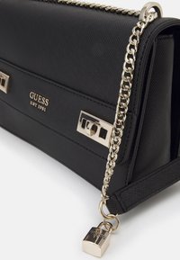 Sort syntetisk håndtaske med en tekstureret overflade, guldfarvede detaljer, kædetras og en hængelåseffekt. Har "GUESS EST 1981" logo.