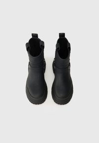 Para czarnych skórzanych botków za kostkę z grubymi podeszwami i uchwytami, widok z góry na jasnym tle.