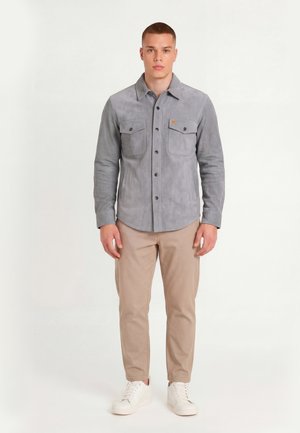 Hombre con camisa gris abotonada con bolsillos en el pecho, pantalones beige y zapatillas blancas, de pie frente a un fondo blanco liso.