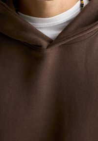 Sweat à capuche marron en mélange de coton. Il présente un col en V, un tissu texturé, et est porté par-dessus un t-shirt blanc.