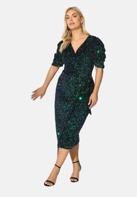 Vihreä ja sininen kimalteleva paljettimekko, jossa on kääntödesign, puffihihat ja polven pituinen helma. Mustat hihnatossut täydentävät asua.