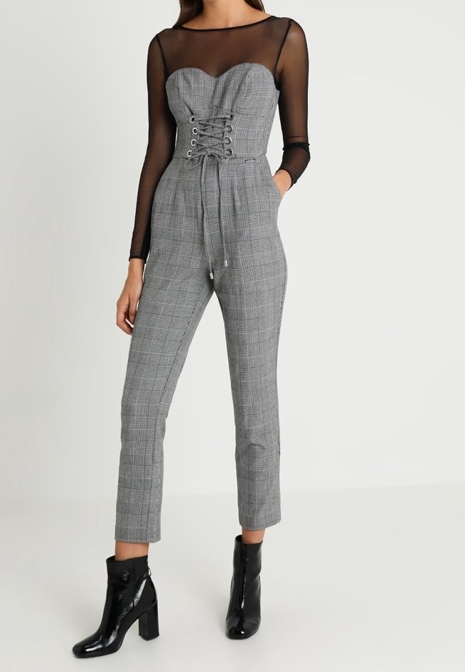 Femme portant une combinaison sans bretelles à carreaux gris avec des manches longues en maille noire et des bottines noires à talons.