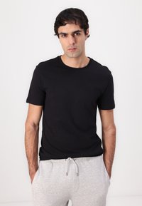 Black crew-neck t-shirt made of cotton, fitted design, short sleeves, worn with gray sweatpants, model standing against a white background.