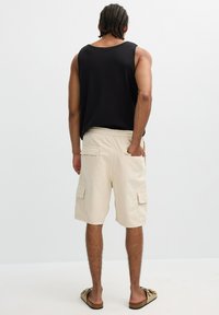 Hombre con trenzas, vestido con una camiseta sin mangas negra, pantalones cortos cargo beige y sandalias marrones, de pie con una mano en el bolsillo trasero.