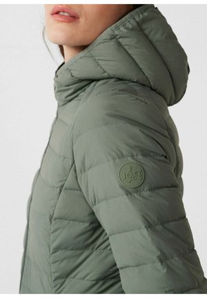 Primo piano di una persona che indossa una giacca imbottita verde oliva con cappuccio e una toppa rotonda sulla parte superiore della manica.