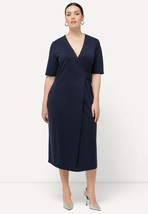 Abito avvolgente blu navy con maniche corte, tessuto morbido, scollatura a V profonda, dettaglio con fiocco laterale, orlo a ginocchio, abbinato a tacchi argentati.