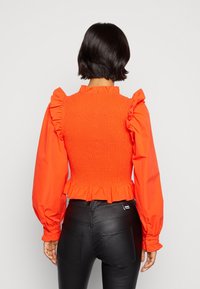 Blusa rossa con schiena arricciata, scollatura a volant e maniche a sbuffo, abbinata a pantaloni neri aderenti. Design texture e vivace.