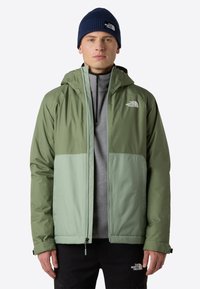 Vihreä vedenpitävä takki, jossa on vetoketjullinen etuosa, kontrastiväriset paneelit ja huppu. Päällä harmaa aluskerros ja mustat housut, kruunattuna sinisellä pipoilla.