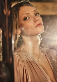 Zlatni nakit sadrži privjesak u obliku sunčevog zraka i usklađene naušnice. Satenska tkanina nježno pada u tonovima blago ružičaste boje.