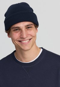 Giovane uomo che indossa un berretto navy e una felpa, sorridendo con uno sfondo chiaro.