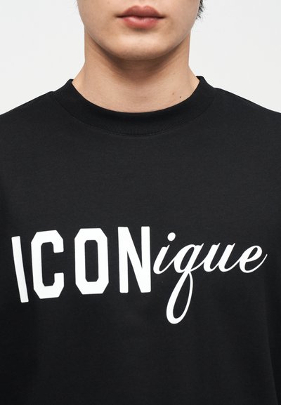 Άτομο που φοράει μαύρο φούτερ με λευκό κείμενο σε ανάμεικτες γραμματοσειρές που γράφει "ICONique" στο στήθος, σε απλό φόντο.