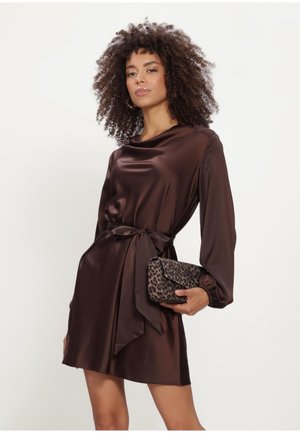 Vrouw met krullend haar, gekleed in een donkerbruine satijnen jurk met lange mouwen, gestrikt in de taille, en een luipaardprint clutch vasthoudend.