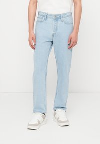 Άνδρας που φοράει τζιν ίσιας γραμμής ανοιχτού μπλε χρώματος και λευκά αθλητικά παπούτσια, στέκεται μπροστά σε απλό λευκό φόντο.