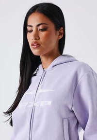 Giovane donna con lunghi capelli neri che indossa una felpa leggera viola con zip e un logo bianco PX, che guarda in basso su uno sfondo semplice.