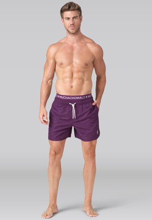 Svalnatý muž stojaci naboso, oblečený v fialových plavkových šortkách s bielym sťahovacím šnúrkou a značkovým elastickým pásom na sivom pozadí.