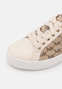Baskets beiges avec lacets blancs, semelle en caoutchouc blanche et panneau latéral en tissu présentant un motif répétitif du logo "MK".