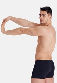 Pantaloncini da bagno neri con una texture liscia, lunghezza fino a metà coscia e un design semplice. Il modello si sta allungando con le braccia distese.
