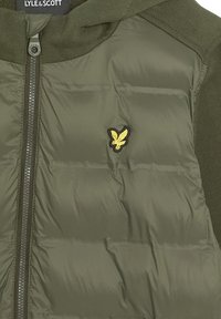 Olivgrüne gepolsterte Jacke mit gestrickten Schultern und Ärmeln, Frontreißverschluss und kleinem gelben Adler-Logo auf der Brust.