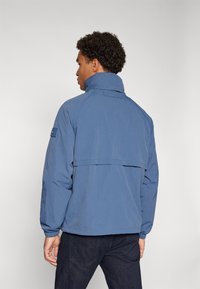 Blue lightweight jacket with a high collar, elastic cuffs, and a back flap design. Features a logo patch on the sleeve.