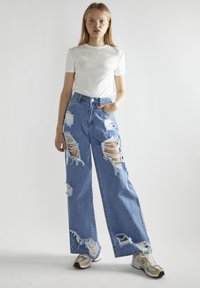 Jeans in denim chiaro e rovinato con grandi strappi, vita alta e silhouette a gamba ampia. Abbinati a una camicia bianca a maniche corte e sneaker.