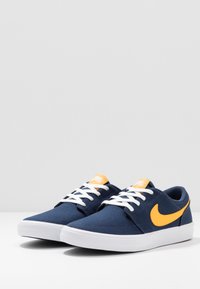 Navy siniset kangaskengät valkoisilla nauhoilla, oransseilla yksityiskohdilla ja valkoisella kumipohjalla. Sivulla on näkyvä oranssi Nike-logo.