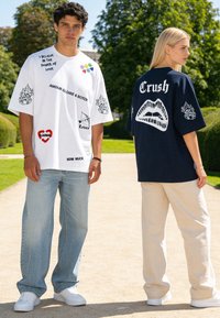 Homme en t-shirt graphique blanc oversize et jeans face à la caméra ; femme en t-shirt graphique bleu marine oversize et pantalon clair debout de profil dans un parc.