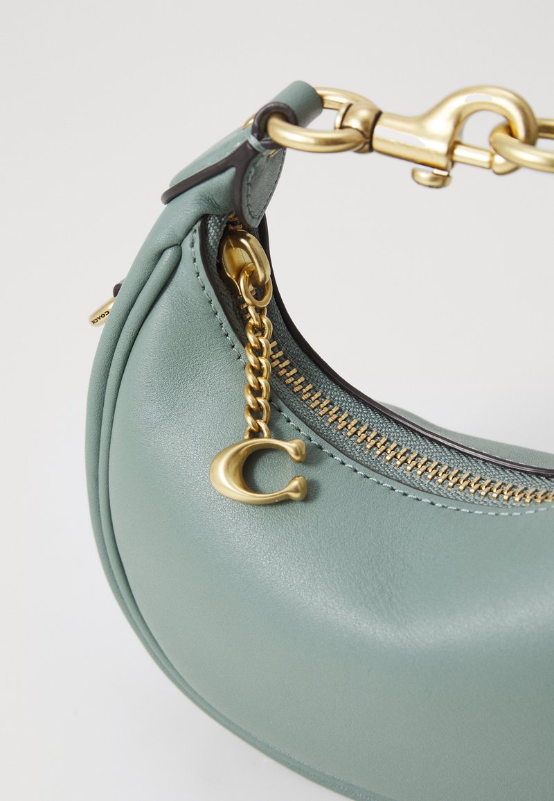 Bolso de mano de cuero verde con forma curva, herrajes en dorado, zipper y un detalle de charm. Textura suave con costuras visibles.