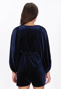 Tengerészkék bársony overál puffos ujjakkal és redőzött derékkal. A hátoldal sima kivitelű, a deréknál rugalmas részlettel.