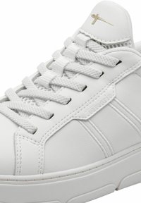 Sneaker en cuir blanc avec des lacets plats blancs, des accents texturés et un logo doré subtil sur la languette. Surface lisse et design minimal.
