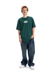 Ciemnozielona, oversize'owa koszulka z okrągłym dekoltem i białym nadrukowanym logo na piersi, w połączeniu z luźnymi niebieskimi dżinsami i białymi butami.