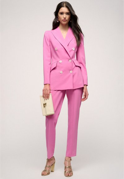 Pink double-breasted suit with notch lapels, button accents, and side pockets. Paired with gold strappy heels and a cream clutch bag.