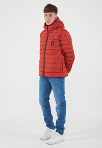 Giacca imbottita rossa con cappuccio, chiusura a zip e tasca sul petto. Indossata con jeans blu e sneakers bianche su uno sfondo neutro.