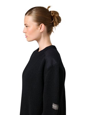 Junge Frau mit Dutt, die einen schwarzen Strickpullover mit einem Aufnäher am Ärmel trägt und nach links vor einem weißen Hintergrund blickt.