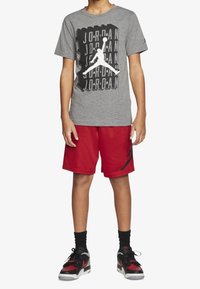 Camiseta gris con logo negro de Jordan, combinada con pantalones cortos rojos, calcetines negros hasta el tobillo y zapatillas deportivas negras/rojas. Diseño atlético simple.