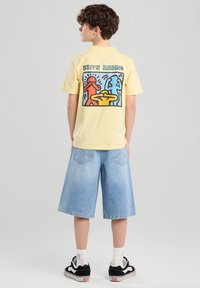 Keltainen puuvillapaita, jossa on Keith Haringin graafinen suunnittelu punaisena, sinisenä ja keltaisena takana. Yhdistetty vaaleansinisiin farkkushortseihin.