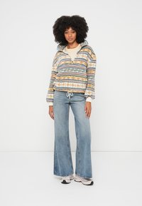 Γυναίκα με σγουρά μαλλιά που φοράει πουλόβερ με ρίγες και ρυθμιζόμενο τελείωμα, φαρδιά τζιν παντελόνια και λευκά αθλητικά παπούτσια, στέκεται μπροστά σε λευκό φόντο.