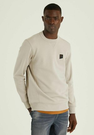 Sudadera de color beige claro con cuello redondo y mangas largas. Presenta un parche cuadrado negro con un logo en el pecho. Tejido de mezcla de algodón.