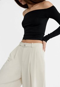 Zwarte off-the-shoulder top van rekbare stof, gecombineerd met losse, lichtbeige plooibroeken met een rechthoekige riemgesp.
