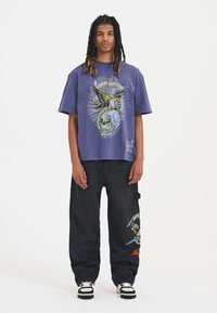 Lilla grafisk t-shirt med et farverigt design af et skelet og en ørn. Kombineret med sorte cargo bukser, der viser et broderet grafisk motiv på benet.