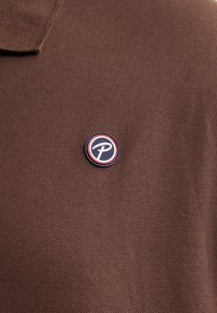 Brązowa koszulka polo z bawełny z okrągłą naszywką logo z niebieską i czerwoną bordiurą oraz stylizowaną literą w środku. Teksturowana wykończenie materiału.