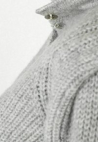Grå stickad tröja med ett texturerat mönster och hög krage, med en detaljerad silverknäppning vid halsen.