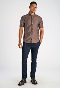Korte mouwen bruin linnen shirt met knoopsluiting, gecombineerd met marineblauwe broek en bruine schoenen, staand op een neutrale achtergrond.