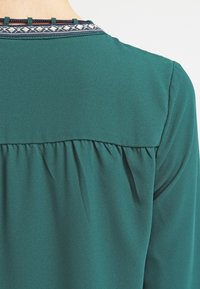 Grön blus med rund ok, med en texturerad halskant i marinblått med ett vitt mönster. Mjuk tyg med rynkdetaljer på baksidan.