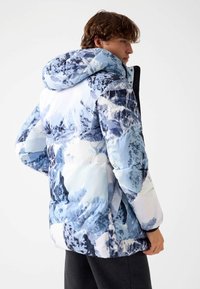 Doudoune avec un imprimé montagne multicolore bleu et blanc, une capuche et une texture matelassée, avec une silhouette ajustée.