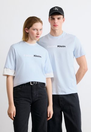 Zwei junge Erwachsene tragen hellblaue Calvin Klein T-Shirts und schwarze Hosen, stehen nebeneinander vor einem schlichten weißen Hintergrund.