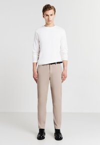 Pantaloni beige su misura con una texture liscia, abbinati a una camicia bianca a maniche lunghe con motivo. Scarpe nere con lacci completano il look.